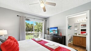A hotel room with a bed, a ceiling fan, a TV, a bathroom with a mirror and lights, and a balcony with a view of palm trees. - Family Getaway Less than 5 miles from Disney! (Kissimmee)