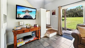 A living room with a TV displaying a beach scene, a wooden console table with books and a DVD player, a leather sofa, and a sliding glass door leading to a grassy outdoor area. - Family Getaway Less than 5 miles from Disney! (Kissimmee)