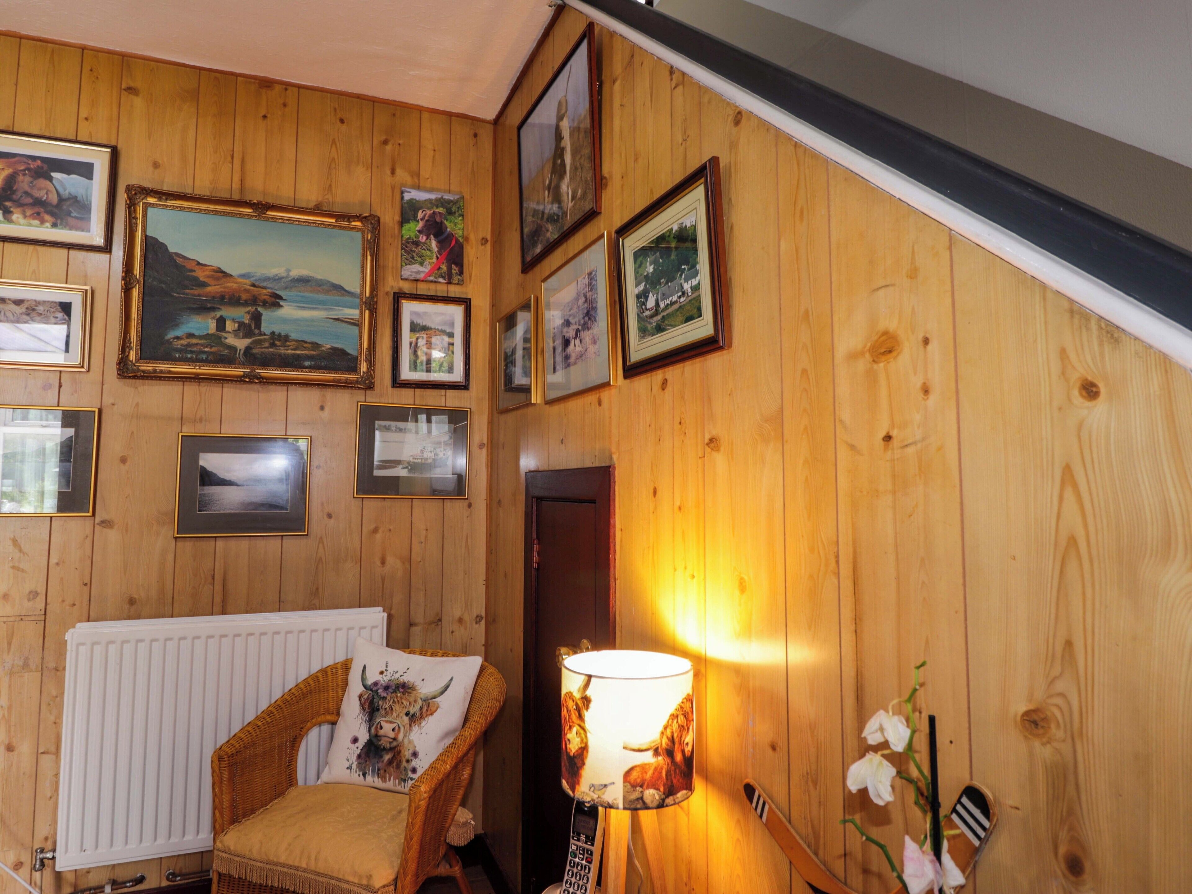 Une pièce chaleureuse avec des murs en bois, un fauteuil en osier, un radiateur et divers tableaux encadrés.