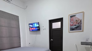 A bedroom with a bed, a TV mounted on the wall, a desk with a chair, and a door. - Shini Guest House (Malang)