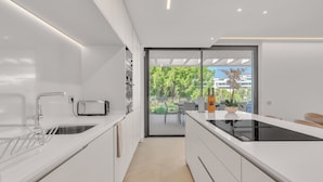 A modern kitchen with white cabinetry, a black induction cooktop, and a large window overlooking a garden. - Villa Soul (Marbella)