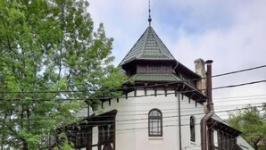 A two-story building with a steeply pitched roof, a tower with a conical roof, and a stone base. There are two cars parked in front, and a tree is visible to the left. - Chateau Sinaia (Sinaia)