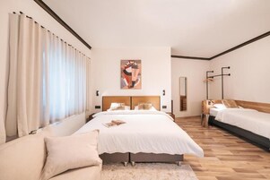 Una habitación de hotel moderna con una cama grande, una cama más pequeña, un sofá y un gran ventanal con cortinas.