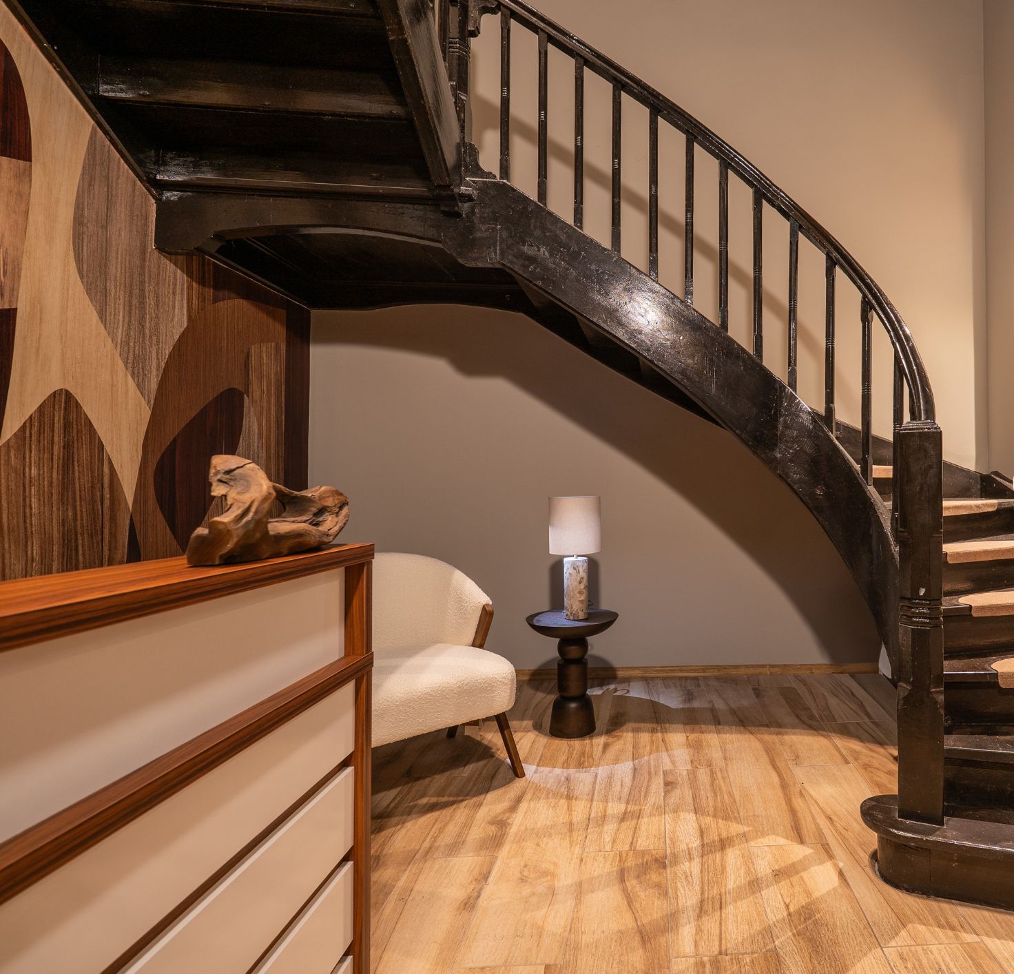 A curved wooden staircase with a black railing, a small round table with a lamp, and a chair in a room with wooden flooring and a beige wall.