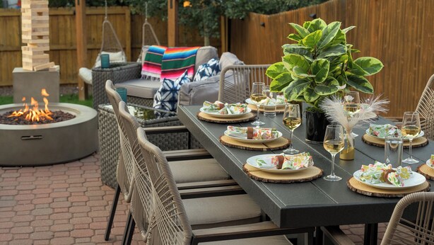 Un espace de restauration en plein air avec une longue table dressée pour un repas, entouré de chaises en osier, d’un foyer et d’une clôture en bois.