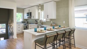 Une cuisine moderne avec des meubles blancs, un îlot central doté de tabourets de bar et une grande fenêtre avec des stores.