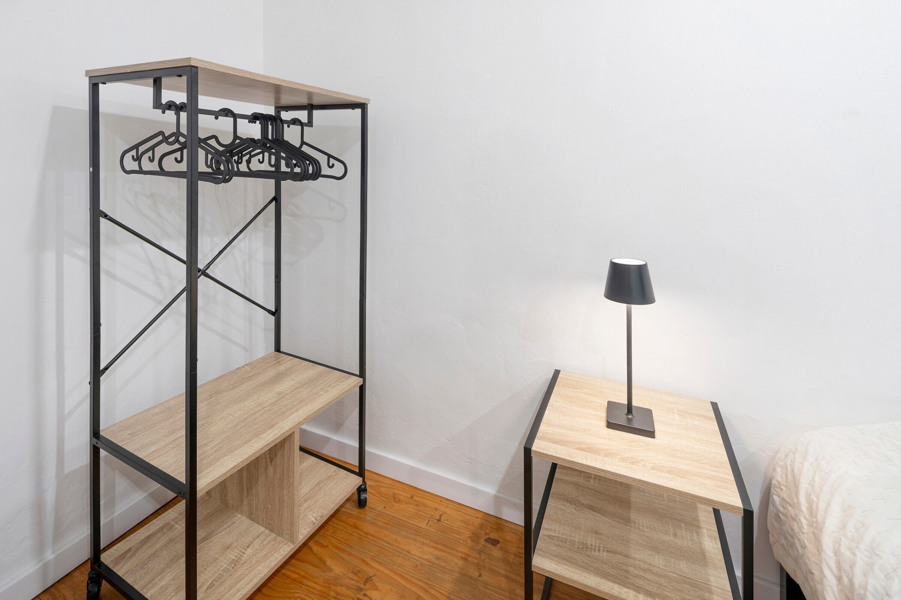 A room with a metal wardrobe, a small wooden table with a lamp, and a bed.