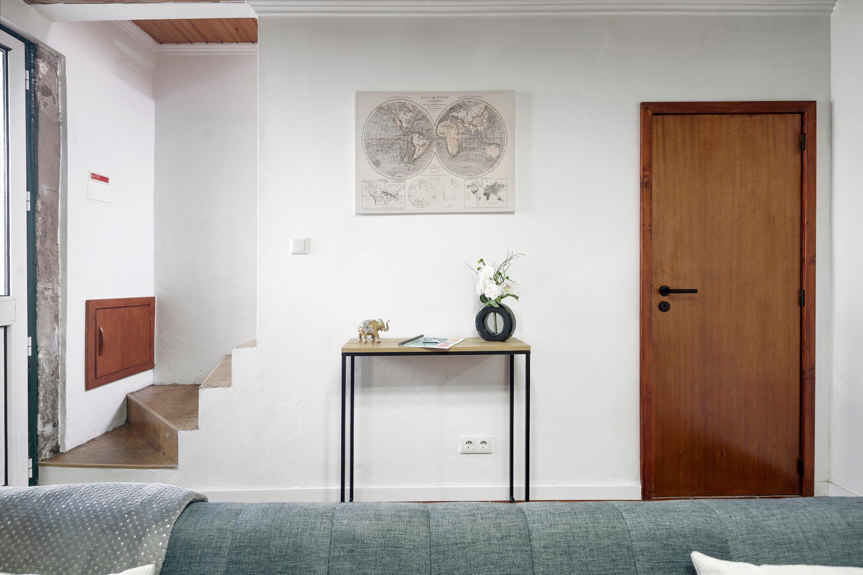 Ein Zimmer mit einer Holztür, einem kleinen Tisch mit Vase und Figur, einer Weltkarte an der Wand und einem grauen Sofa.