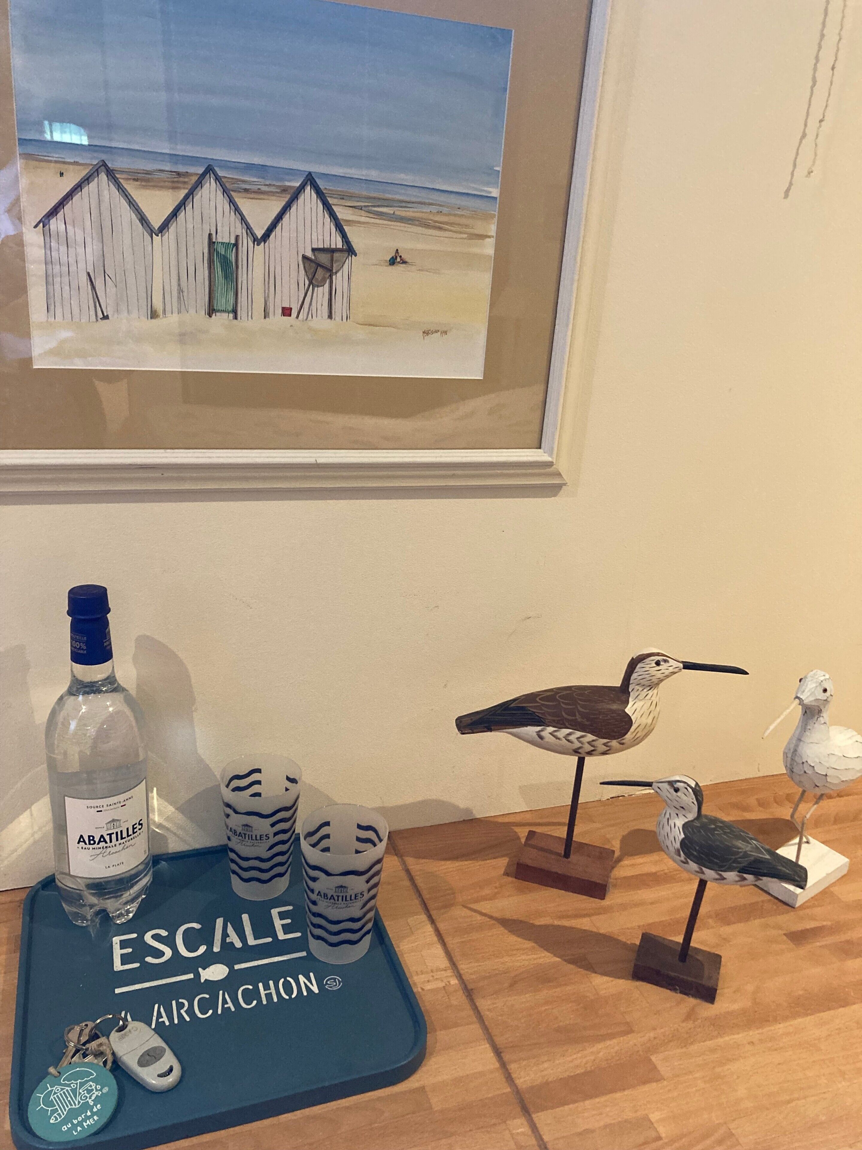Un tableau encadré représentant des cabanes de plage, une bouteille d’Abatilles et des statuettes d’oiseaux disposées sur un plateau.