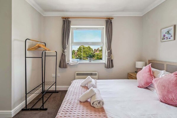 Un dormitorio con cama, ventana con cortinas, radiador, mesita de noche con lámpara y un cuadro colgado en la pared.