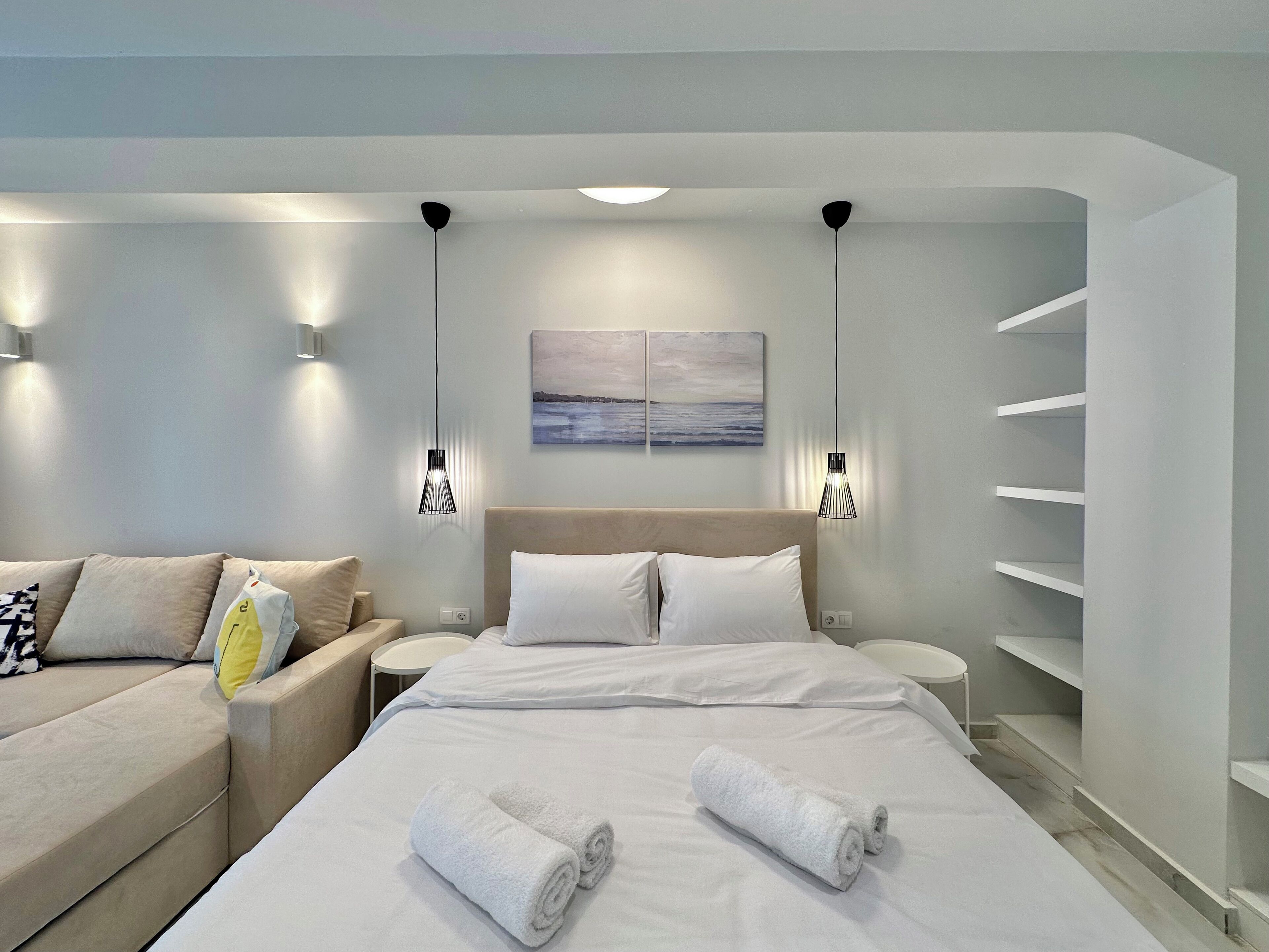A modern hotel room with a large bed, a sofa, and a small table. There are wall-mounted lights and a painting above the bed.