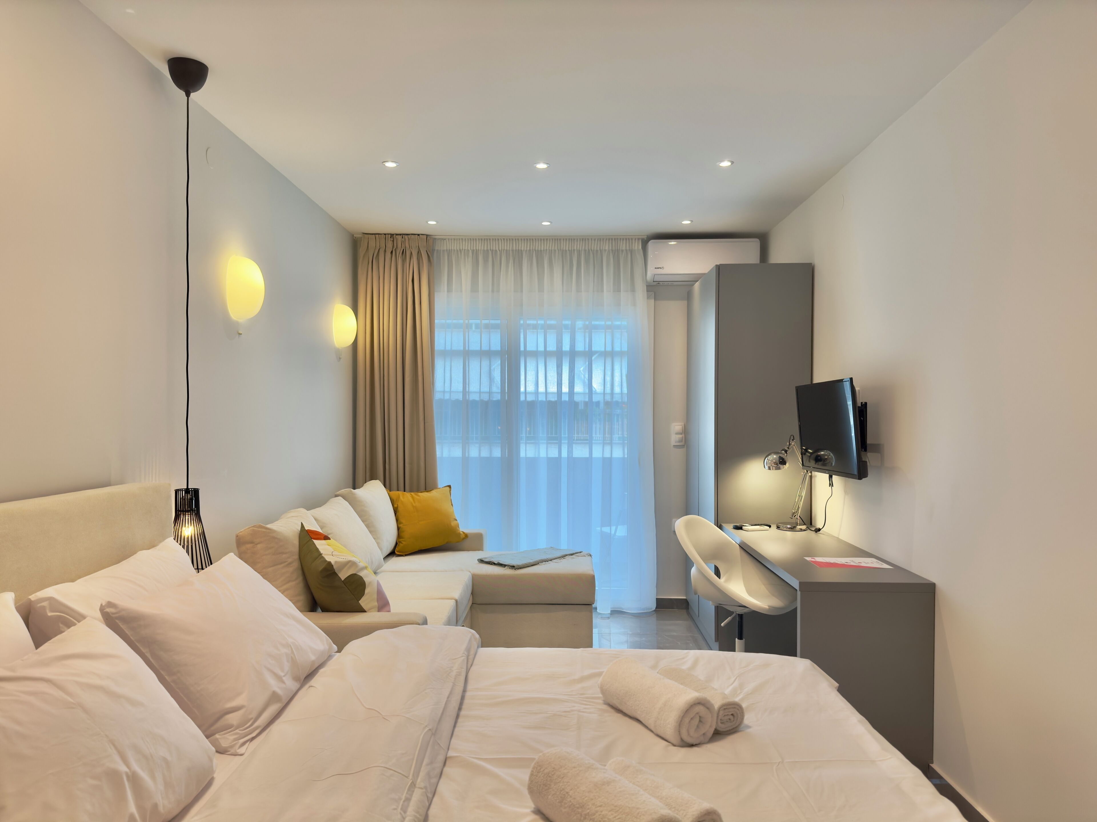 Una habitación de hotel moderna con cama, escritorio, televisor y un amplio ventanal con cortinas.