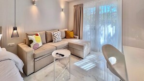 A modern living room with a sectional sofa, a small round table, and a chair. There is a ceiling light fixture and an air conditioning unit.