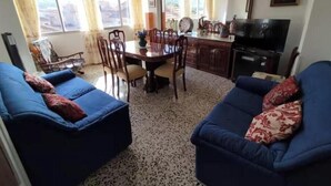 Un salon avec deux canapés bleus, une table à manger avec des chaises, un coin cuisine avec des placards et un téléviseur, ainsi qu’une grande fenêtre avec des rideaux.