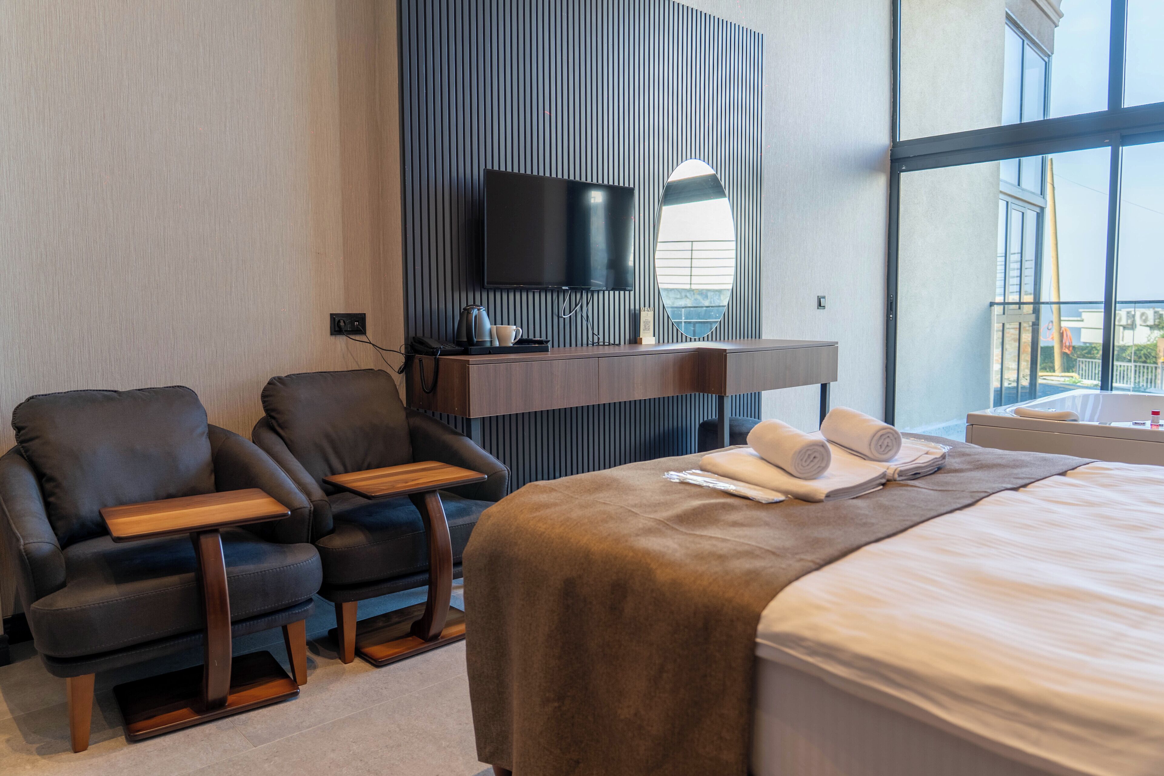 A hotel room with a bed, two armchairs, a TV, and a view of the city.