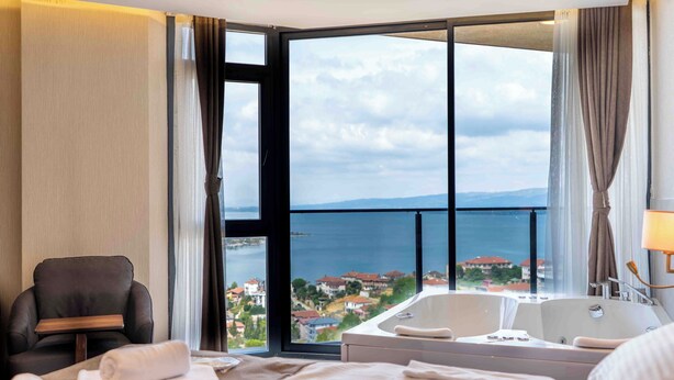 A hotel room with a large window, a bathtub, a bed, a chair, and a table with towels.
