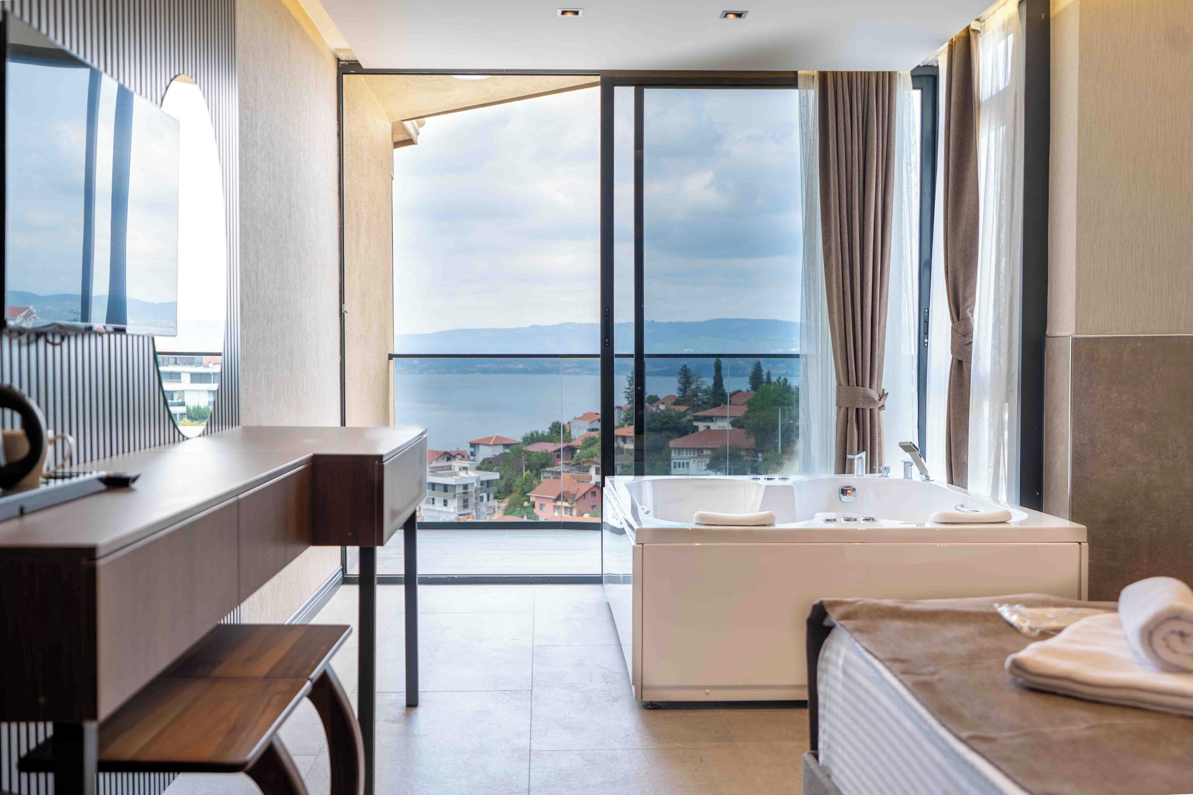A modern hotel room with a large window, a bathtub, a desk, and a view of the sea.