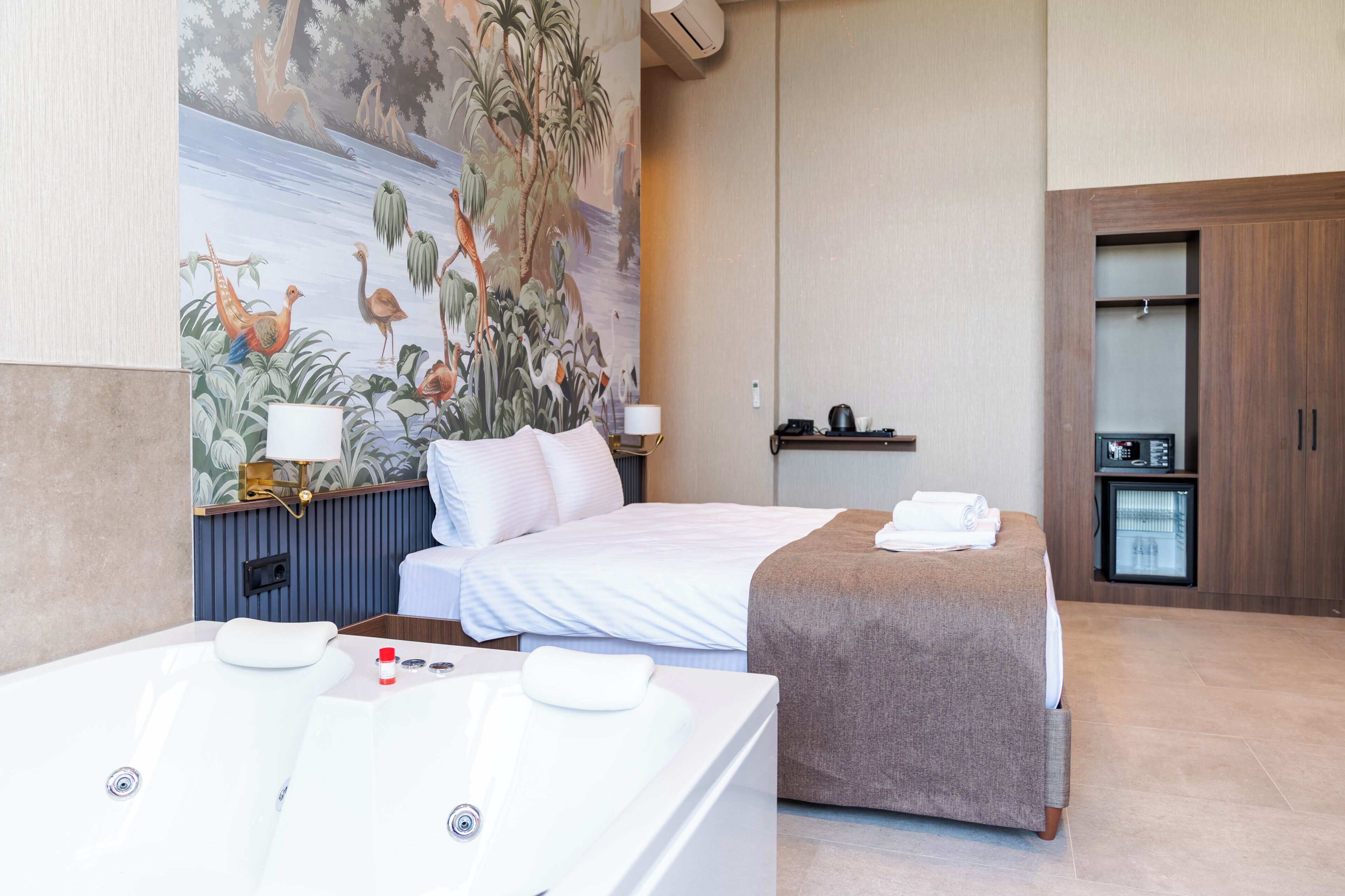 A hotel room with a large mural, a double bed, a separate bed, a bathtub, and a minibar.