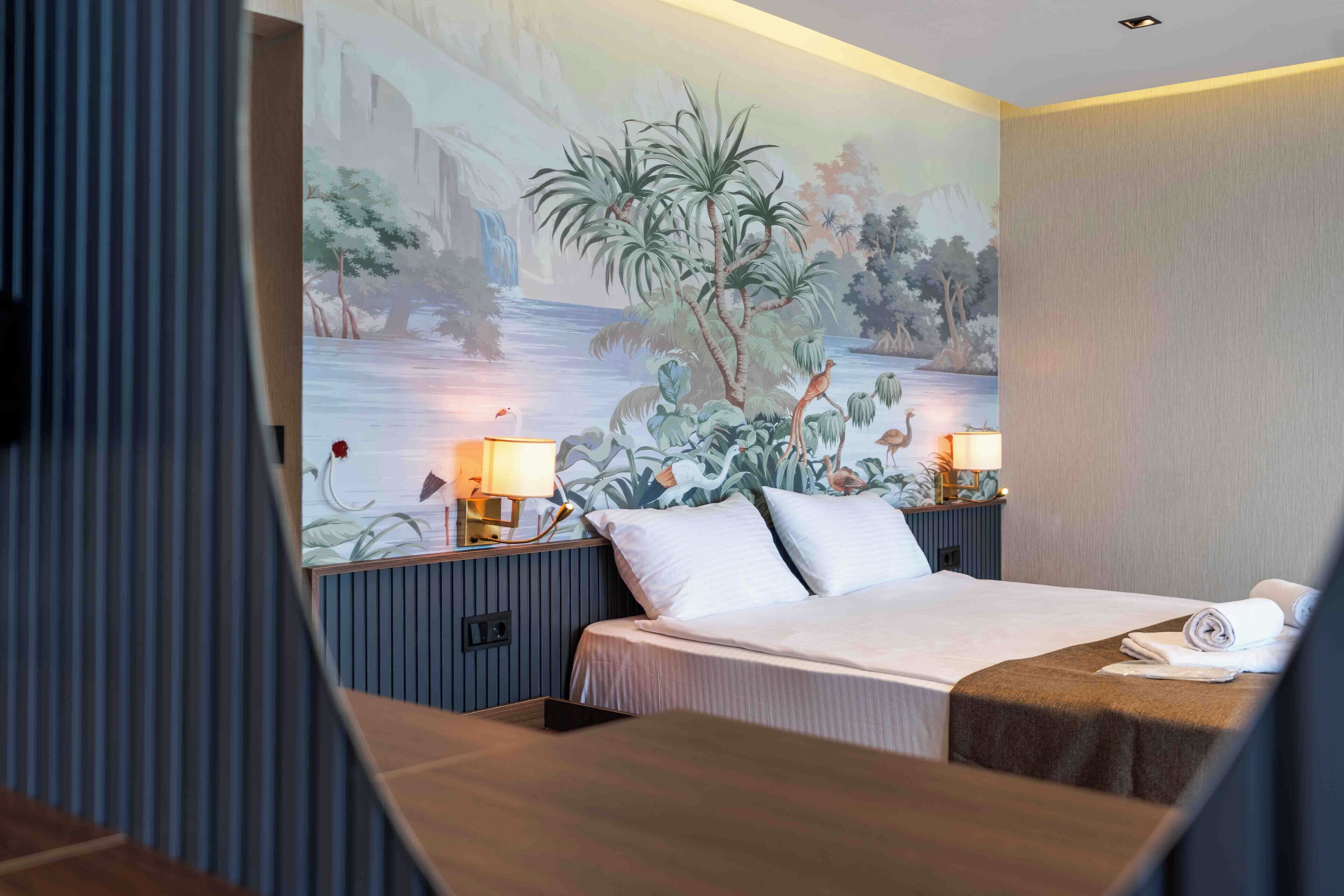 A hotel room with a bed, two bedside tables with lamps, a mural on the wall, and a window with curtains.