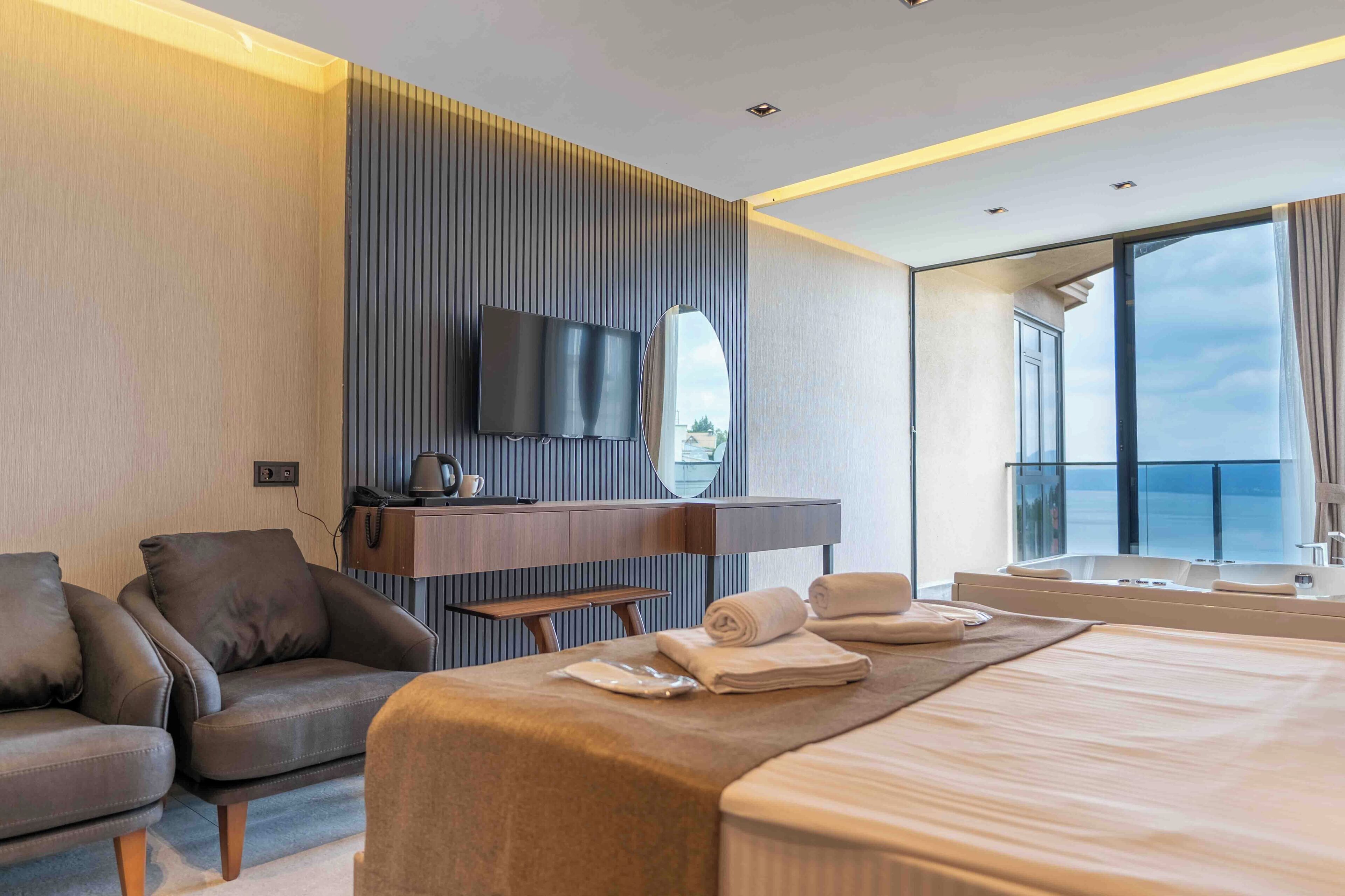 A modern hotel room with a large bed, a seating area with a chair, a flat-screen TV mounted on a wall, and a large window offering a view of the sky.