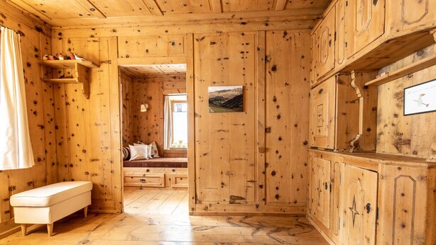 Ein Holzblockhaus im Inneren mit einer Bank, einem kleinen Tisch und einem Fenster mit Blick auf die Berge.
