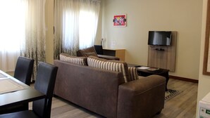 Una sala de estar con un sofá marrón, almohadones a rayas, una mesa de comedor con sillas, un televisor sobre un mueble de madera y una ventana con cortinas traslúcidas.