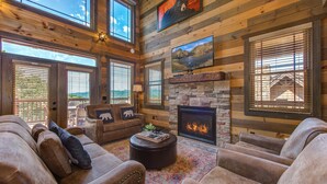 A spacious living room with a stone fireplace, leather sofas, and large windows offering a view of the outdoors.