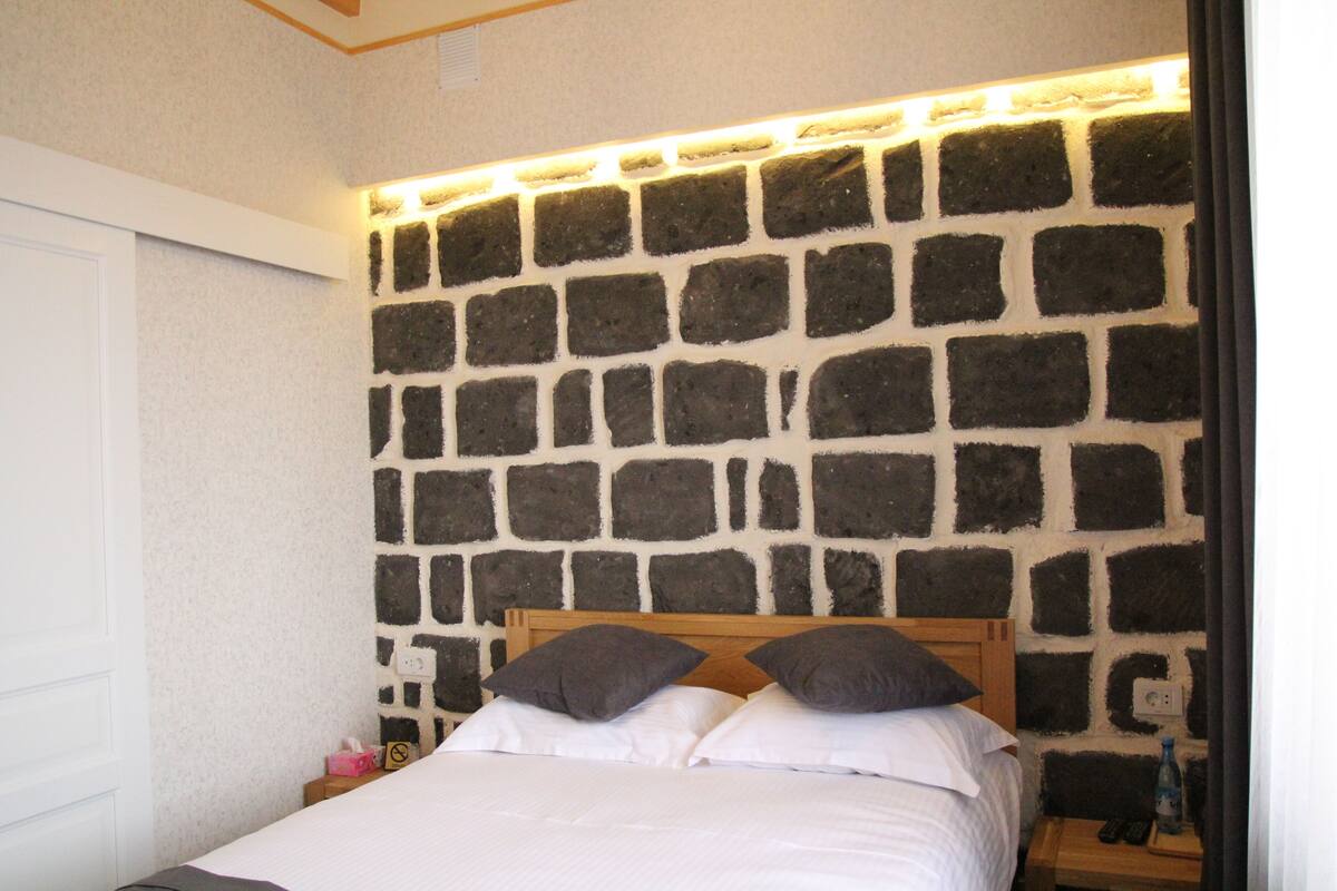 A bedroom with a stone wall headboard, a wooden bed frame, and a bedside table with a remote control and a bottle.