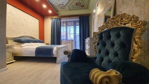 Habitación de hotel con una cama grande, una zona de estar separada con cabecero capitonado, un radiador y una ventana con cortinas.
