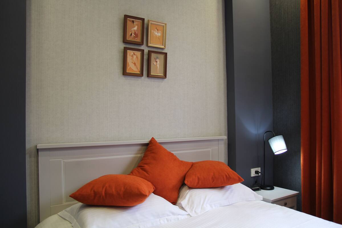 A hotel room with a bed, orange pillows, a nightstand with a lamp, and framed pictures on the wall.