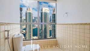 Une salle de bain dotée d’une grande fenêtre donnant sur le paysage urbain de Dubaï, avec ses nombreux gratte-ciel et son port de plaisance.
