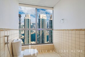 A bathroom with a large window offering a view of Dubai's skyline, including tall buildings and a marina. - LUX The JBR Central Suite (Dubai)
