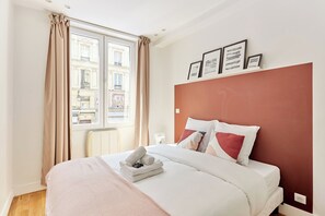 Habitación de hotel con una cama grande, cortinas color beige, una ventana con vistas a edificios y una pared de acento roja.
