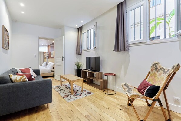A modern living room with a grey sofa, a wooden coffee table, a folding chair, a TV on a wooden stand, and a window with curtains. - Superb Apartment - Beaubourg - Le Marais (Paris)