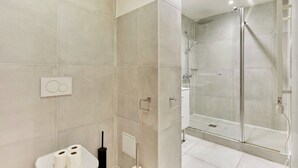 Un baño moderno con baldosas color beige, un inodoro con dos rollos de papel higiénico, un cepillo de inodoro negro y un box de ducha de vidrio.