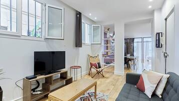 A modern living room with a flat-screen TV, wooden entertainment unit, grey sofa, and a dining area in the background.