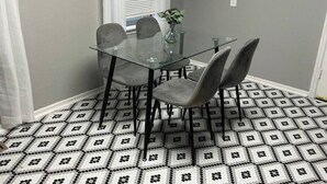 Uma sala de jantar com mesa de vidro e quatro cadeiras, um vaso com planta e um chão estampado.
