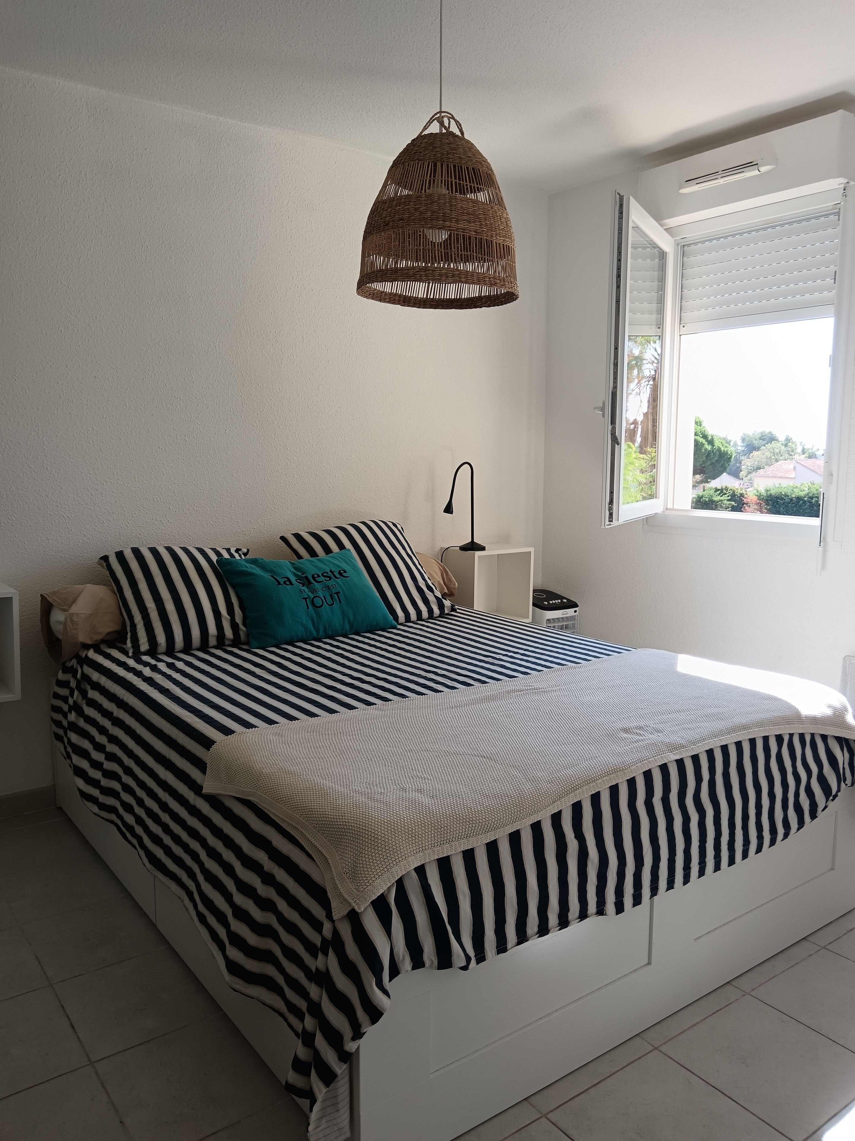 A neatly made bed with striped bedding, a hanging wicker light fixture, a bedside lamp, and a window with blinds.