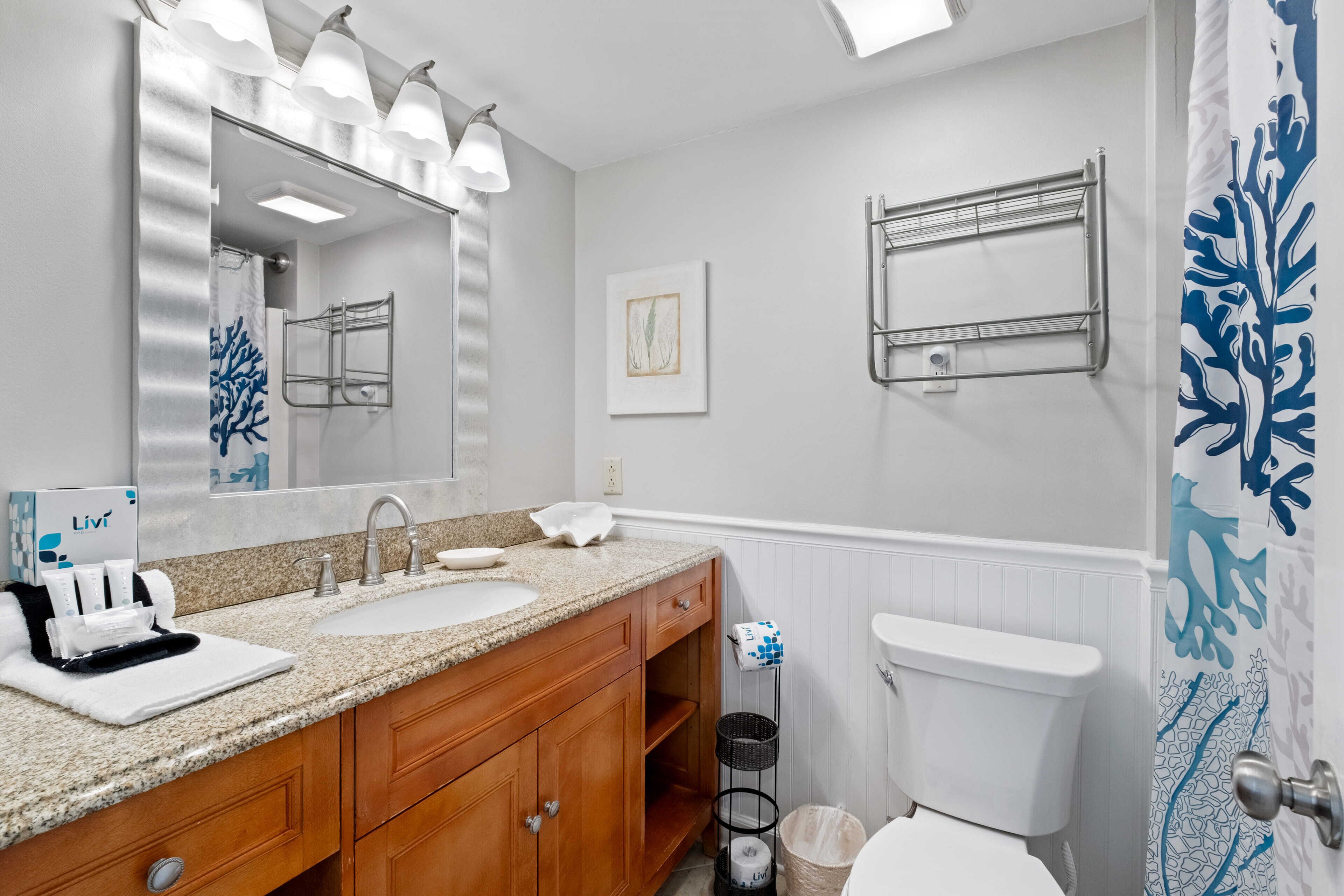 A bathroom with a double sink vanity, a mirror with lights, a towel rack, a toilet, and a shower curtain with a blue coral pattern.