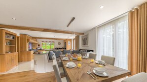 A spacious dining area with a wooden table set for breakfast, a kitchen in the background, and large windows with curtains.