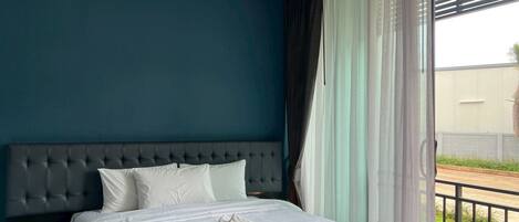 Habitación de hotel con pared azul oscuro, cabecera capitonada, cama grande con ropa de cama blanca y vista a un balcón con barandilla.