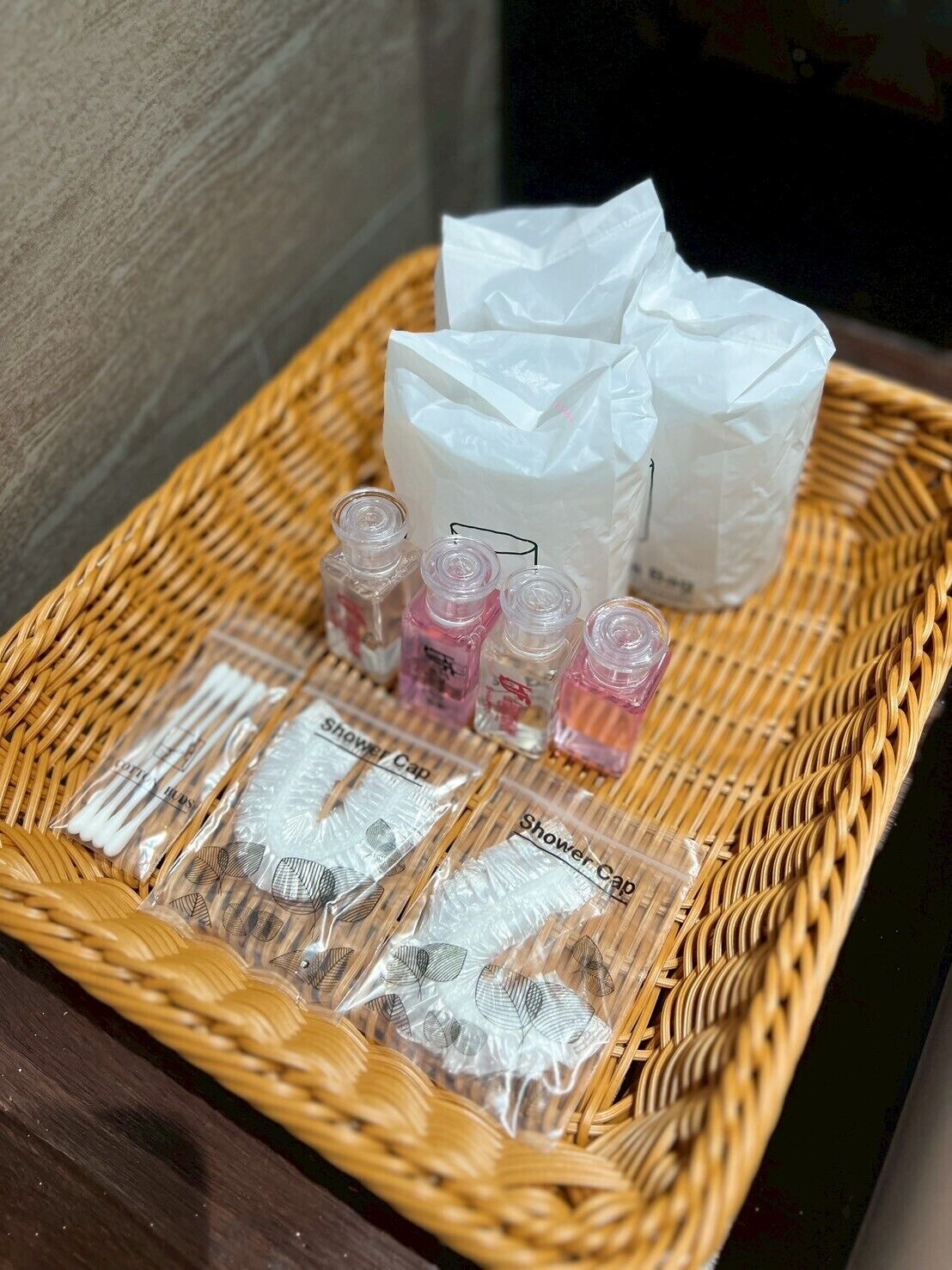 A wicker basket containing shower caps, a white paper bag, and small bottles of liquid soap.