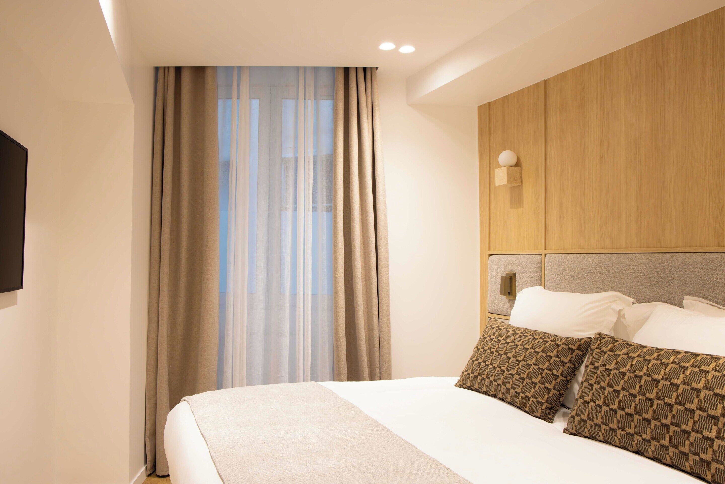 Une chambre d’hôtel avec un grand lit, des rideaux beiges, une tête de lit en bois et un téléviseur fixé au mur.