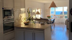 A modern kitchen with white cabinetry, an oven, and a microwave. There is a dining area with a table and chairs, and a view of the ocean through large windows. - Magnificent duplex with garden and sea view - Beach and Sanary on foot (Six-Fours-les-Plages)