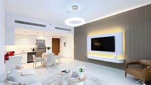 A modern living room with a flat-screen TV mounted on a wall, a sofa, a coffee table, and a dining area with chairs. - Luxury & Comfort 1B at Al Jadeel 2 Jumeirah Living (Dubai)