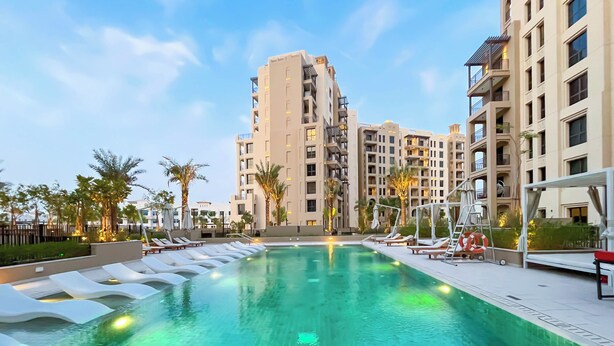 A swimming pool with lounge chairs, surrounded by palm trees and modern buildings. - Luxury & Comfort 1B at Al Jadeel 2 Jumeirah Living (Dubai)