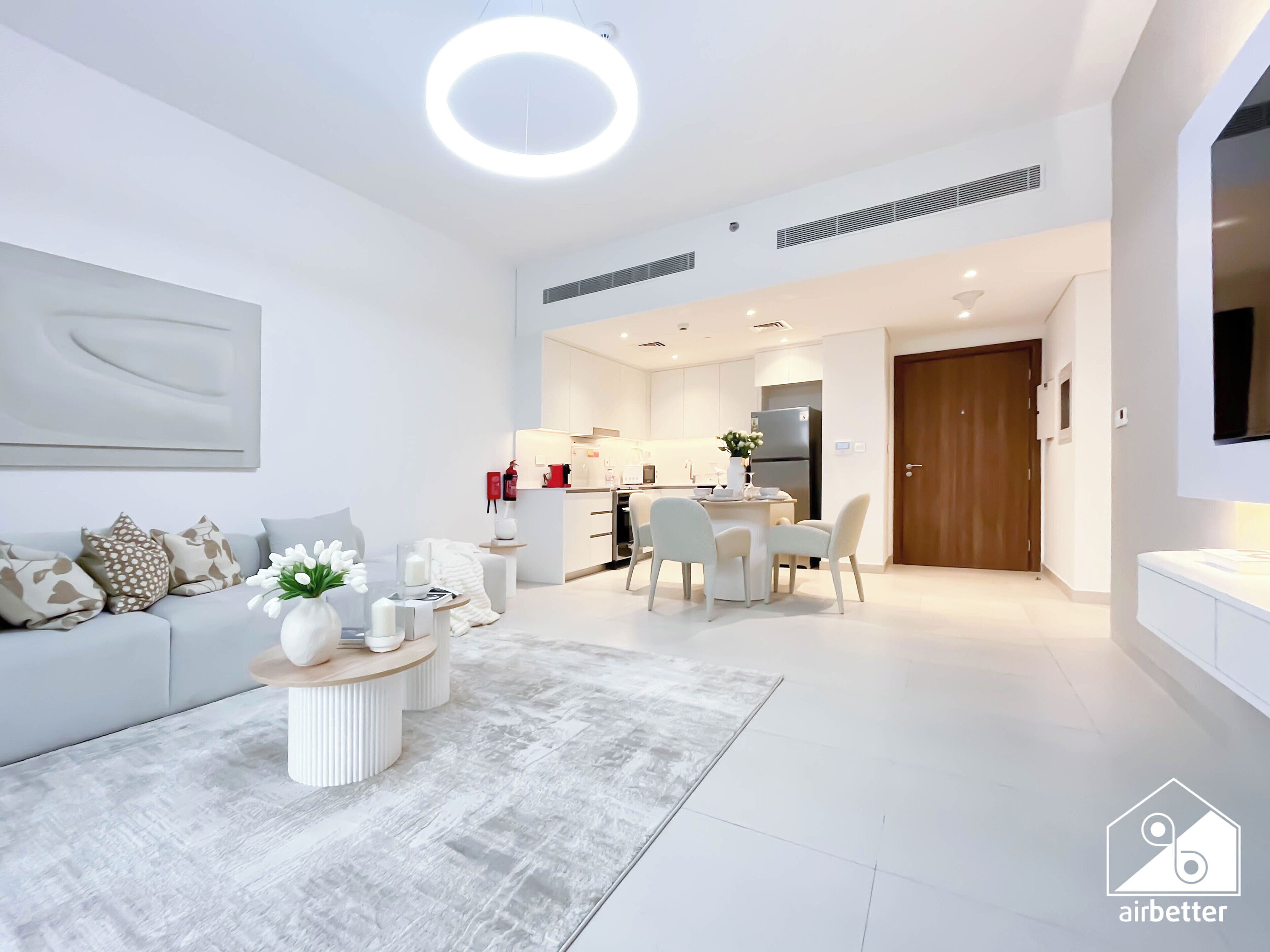 Ein modernes Wohnzimmer mit Sofa, Couchtisch und Essbereich mit Stühlen. Im Hintergrund ist eine Küche mit Kühlschrank und Mikrowelle erkennbar.