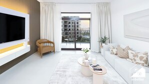 A modern living room with a flat-screen TV, a comfortable chair, a sofa with patterned cushions, a coffee table, and a view of a cityscape through large windows. - Luxury & Comfort 1B at Al Jadeel 2 Jumeirah Living (Dubai)