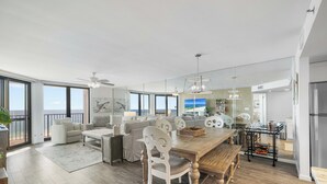 A spacious living area with a wooden dining table, white chairs, and a large window overlooking the ocean.