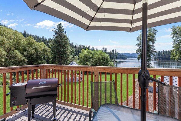 A deck with a grill, a chair, and a view of a lake and forest. - New Listing! Hayden Lake Guest House | Private Balcony & Lake Views (Hayden)